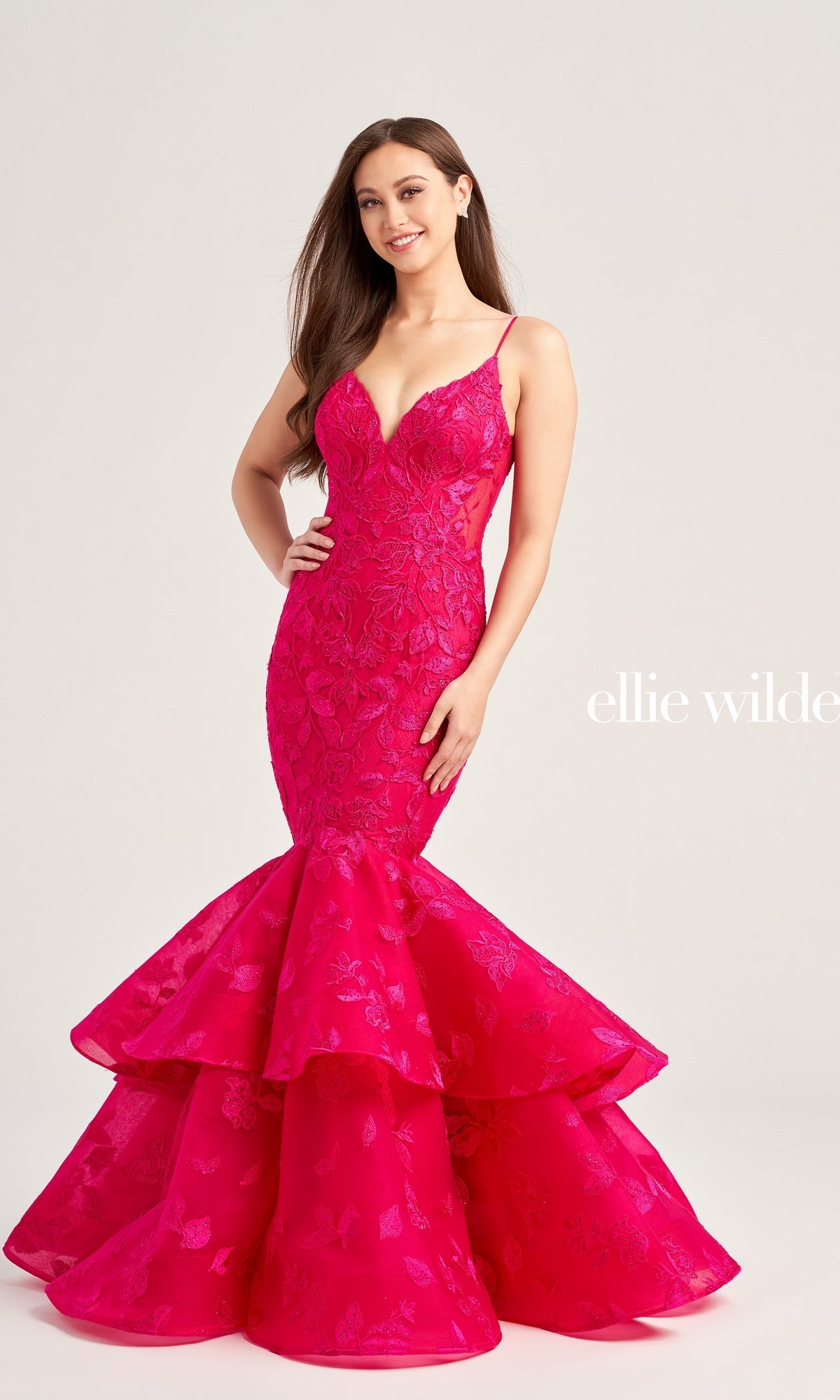 A woman wears the Ellie Wilde Long Lace Mermaid Prom Dress EW35092 in bright pink floral lace, featuring spaghetti straps and a ruffled flared hem, standing against a plain light background.